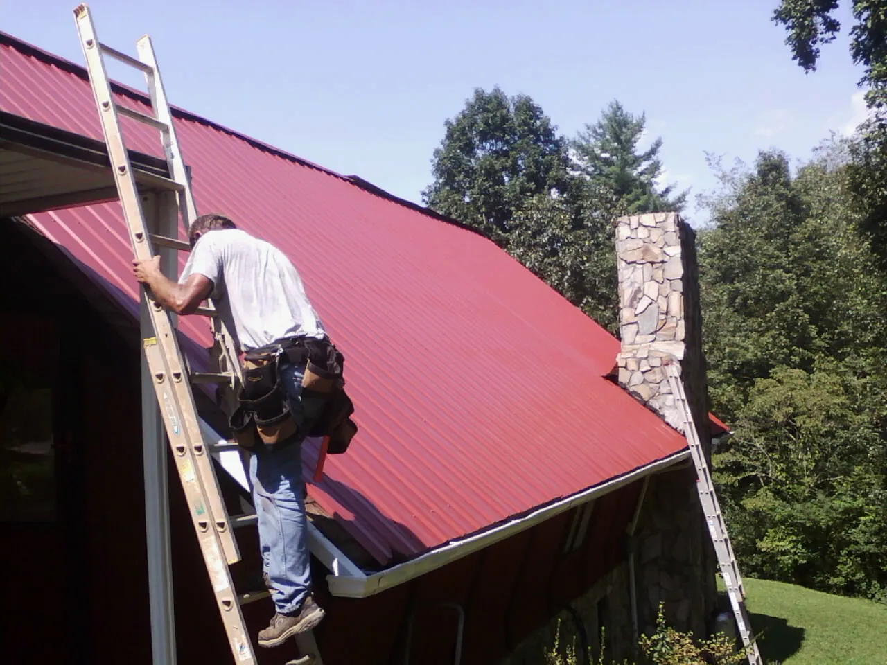Roofer on a ladder beside a red metal roof and stone chimney — Soffit & Fascia Repair by Roofing Brothers in Sandpoint
